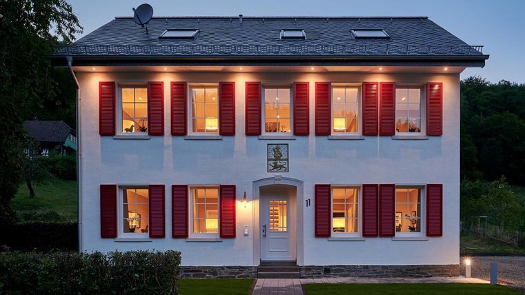 Ein modernes, zweistöckiges Haus mit weißen Wänden und roten Fensterläden. Die Fenster sind beleuchtet und es gibt einen schmalen Weg zur Eingangstür.