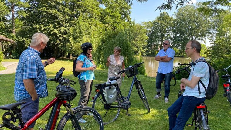 Eine Gruppe von sechs Personen steht mit Fahrrädern auf einer grünen Wiese. Im Hintergrund sind Bäume und ein kleiner Teich zu sehen.