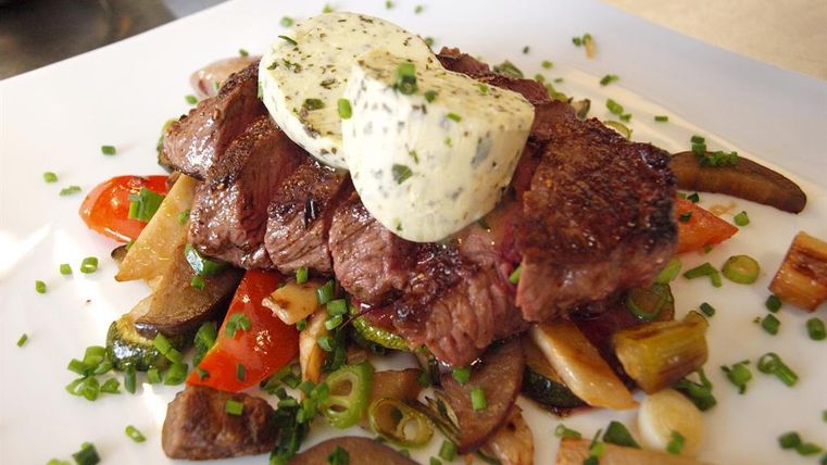 Ein saftiges Stück Rindersteak auf einem Bett aus Gemüse. Darauf ist ein Stück Kräuterbutter angerichtet.