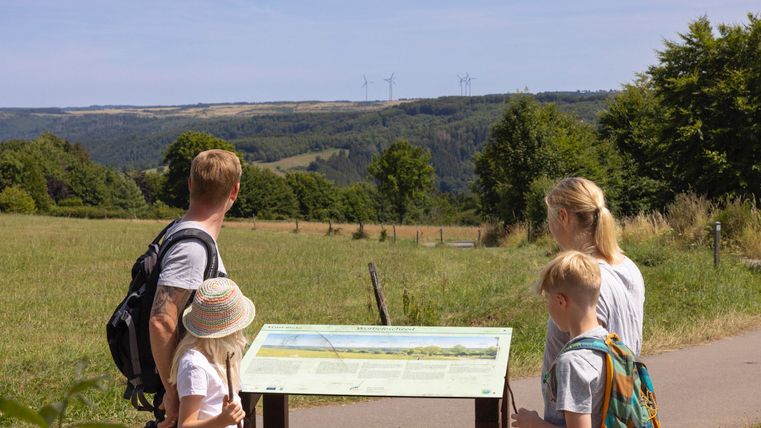Eine Familie steht vor einem Informationsschild in einer grünen Landschaft. Im Hintergrund sieht man hügelige Felder und Bäume unter einem klaren Himmel.