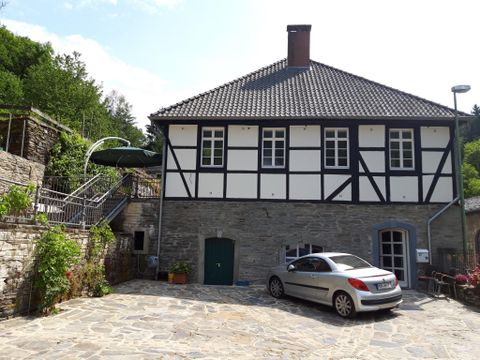 Ein historisches Haus mit Fachwerkstil und einem gepflasterten Hof. Im Vordergrund steht ein graues Auto, während der Garten im Hintergrund grün ist.