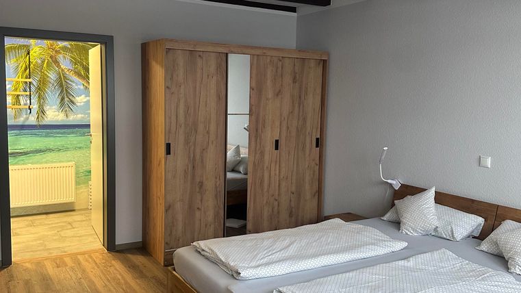 Ein modernes Schlafzimmer mit einem großen Doppelbett und einem Kleiderschrank aus Holz. Im Hintergrund ist ein Fenster mit Blick auf eine sonnige Landschaft.
