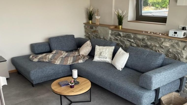 Ein modernes Wohnzimmer mit einem grauen Sofa und einem runden Holztisch. Im Hintergrund befindet sich eine Steinwand und eine große Fensterfront.