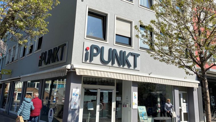Ein modernes Geschäft mit dem Namen "iPUNKT" in einer städtischen Umgebung. Menschen gehen vorbei und es gibt Bäume, die Schatten spenden.
