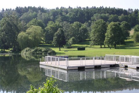Ein ruhiger See mit einer Holzplattform im Vordergrund. Im Hintergrund sind grüne Bäume und sanfte Hügel zu sehen.