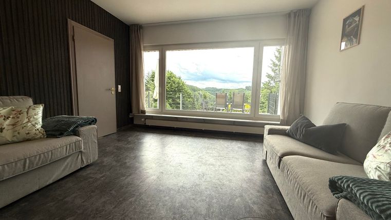 Ein modernes Wohnzimmer mit großen Fenstern und Blick auf die Natur. Die Möbel sind einfach und stilvoll gestaltet.