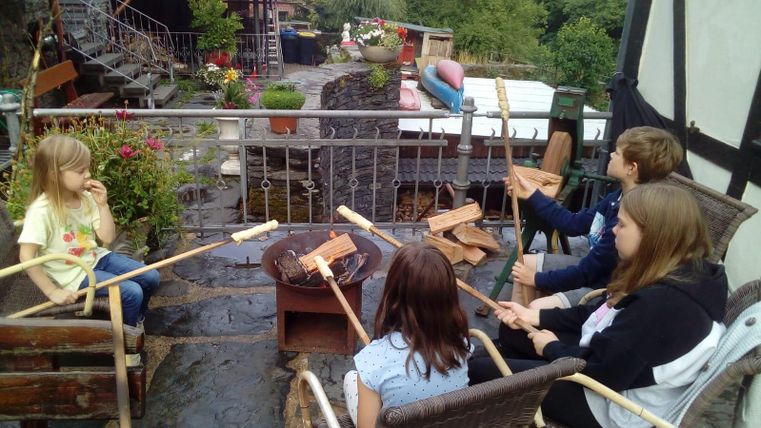 Eine Gruppe von Kindern sitzt um ein Feuer, während sie Stockbrot zubereiten. Im Hintergrund sind Blumen und eine rustikale Landschaft zu sehen.