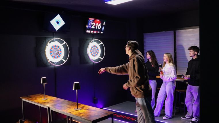Eine Person spielt Darts in einem modernen Raum mit digitalen Zielscheiben. Im Hintergrund stehen drei weitere Personen und beobachten das Spiel.
