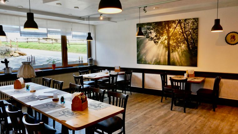 Ein gemütliches Restaurant mit Holztischen und Stühlen. An der Wand hängt ein großes Bild mit Sonnenstrahlen, die durch Bäume scheinen.
