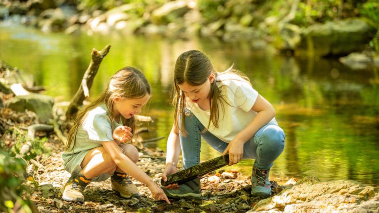Zwei Mädchen spielen am Ufer eines kleinen Bachs. Sie untersuchen gemeinsam einen Stock im Wasser.