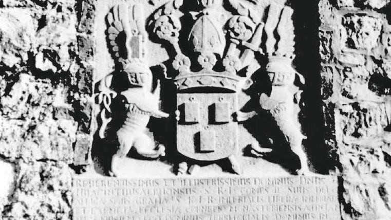 Ein Wappen mit einem königlichen Schild und zwei Löwen, die es halten. Der Hintergrund besteht aus einer Steinwand.