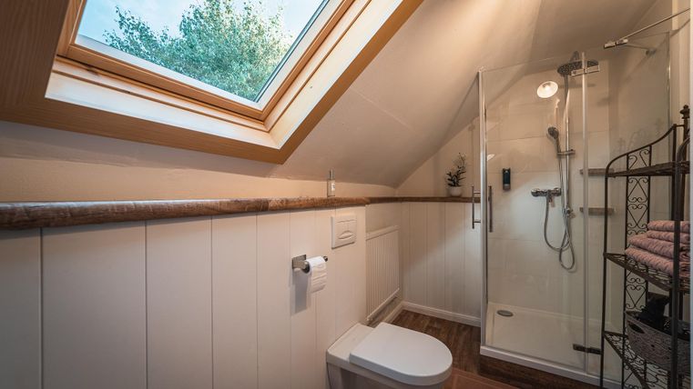 Ein modernes Badezimmer mit einer Dusche und einem Fenster, das Tageslicht hereinlässt. Es gibt auch eine Toilette und einen Regalständer mit Handtüchern.
