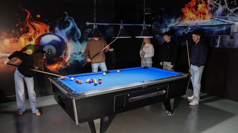 Eine Gruppe von vier Personen spielt Billard an einem blauen Tisch. Im Hintergrund sind farbenfrohe, dynamische Grafiken zu sehen.