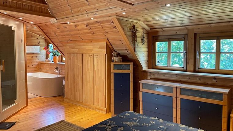 Ein gemütliches Schlafzimmer mit Holzverkleidung und Fenstern, die viel Licht hereinlassen. Im Hintergrund ist ein modernes Badezimmer mit Badewanne zu sehen.