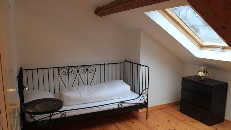 Ein gemütliches Zimmer mit einem Eisenbett, einem kleinen Tisch und einer Kommode. Das Licht fällt durch ein Fenster in der Dachschräge.