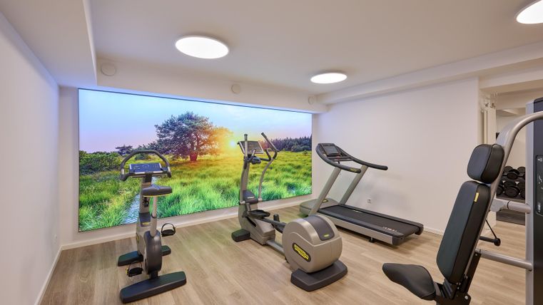 Ein moderner Fitnessraum mit verschiedenen Trainingsgeräten. Im Hintergrund ist ein großes Bild einer grünen Landschaft mit Sonnenaufgang zu sehen.