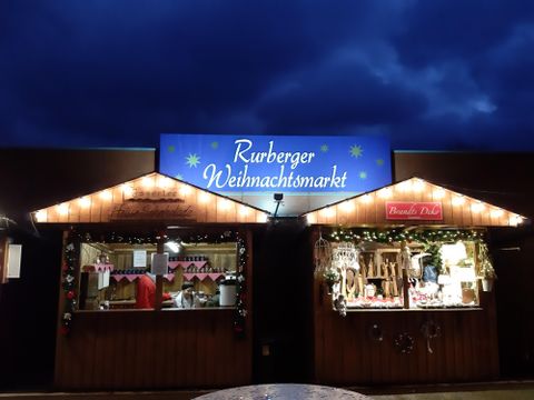 Ein Weihnachtsmarkt mit zwei festlich dekorierten Ständen. Der Himmel ist dunkel und es leuchten Lichter an den Ständen.