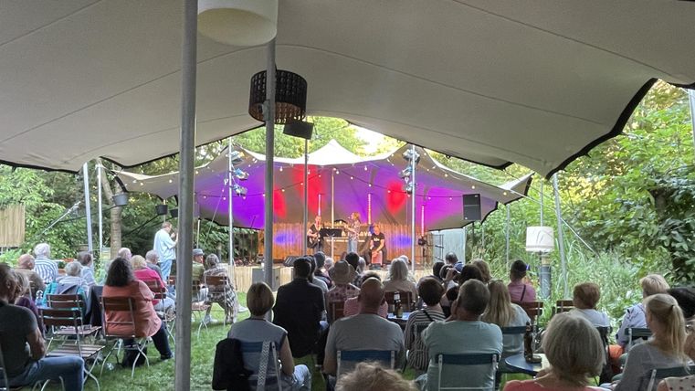 Ein Konzert unter einem großen Zelt mit vielen Zuschauern. Buntes Licht erhellt die Bühne, während Musiker spielen.
