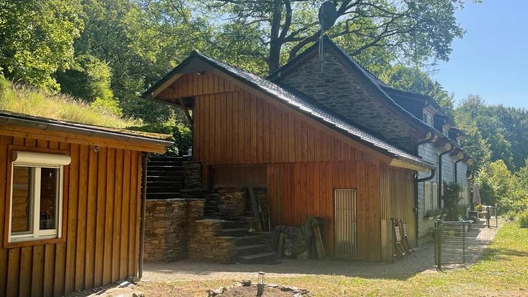 Ein Holzgebäude in einer grünen Umgebung mit Bäumen im Hintergrund. Der Garten ist unordentlich mit einem kleinen Steinhaufen.