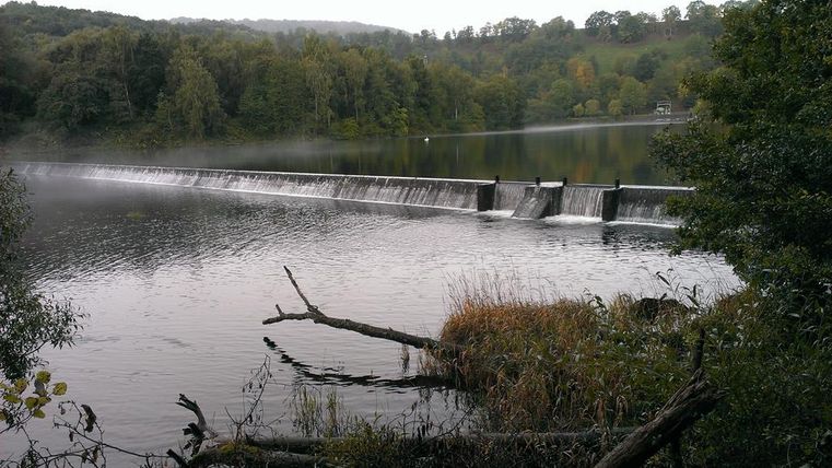 Ein Staudamm erstreckt sich über einen ruhigen See, umgeben von Bäumen und sanften Hügeln. Der Wasserfall fließt sanft über die Kante des Damms.