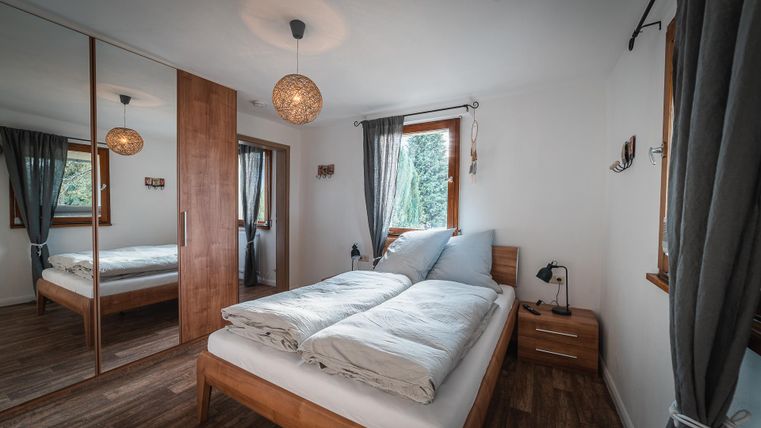 Ein gemütliches Schlafzimmer mit einem Doppelbett und Holzmöbeln. Große Fenster sorgen für viel Tageslicht und eine entspannte Atmosphäre.