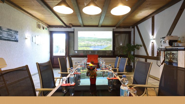 Ein modernes Konferenzzimmer mit einem langen Tisch und Stühlen. An der Wand befindet sich ein Projektor, und das Fenster bietet Blick ins Freie.