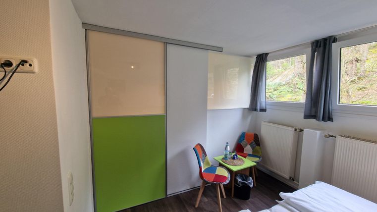 Ein modernes Zimmer mit einem grünen Schrank, einem kleinen Tisch und bunten Stühlen. Die Fenster bieten Blick auf die Natur.