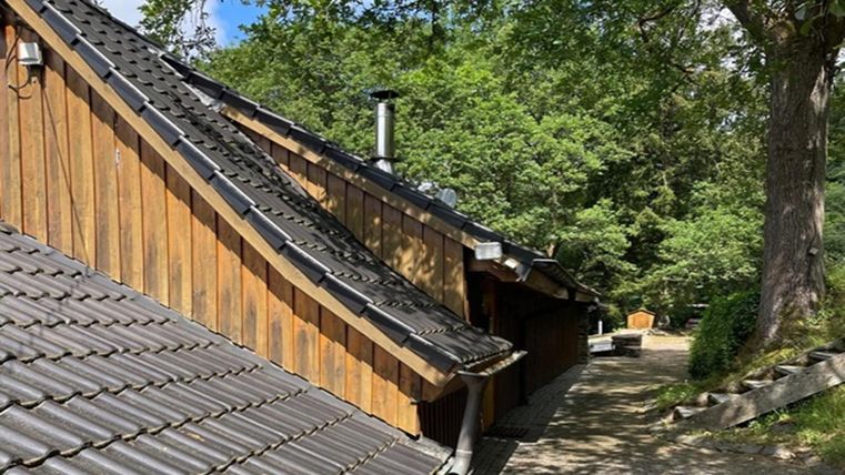 Ein Blick auf ein Holzhaus mit einem schrägen Dach und grünen Bäumen im Hintergrund. Der Weg neben dem Haus ist von Natur umgeben.