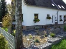 Ein hübsches Haus mit weißen Wänden und einem Garten aus Kies. Bunte Blumenstöcke zieren die Fenster.