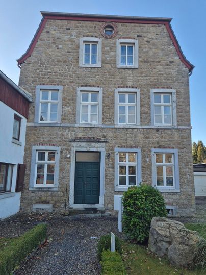 Ein historisches Steinhaus mit einer grünen Eingangstür und großen Fenstern. Umgeben von einem kleinen Garten und ein paar Steinen im Vordergrund.