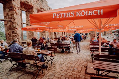 Eine einladende Freiluftterrasse mit Tischen und Stühlen unter orangefarbenen Schirmen. Gäste genießen Getränke in einer rustikalen Umgebung.