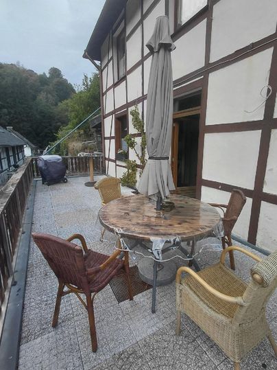 Eine Terrasse mit einem runden Holztisch und mehreren Stühlen. Im Hintergrund ist ein traditionelles Fachwerkhaus zu sehen.