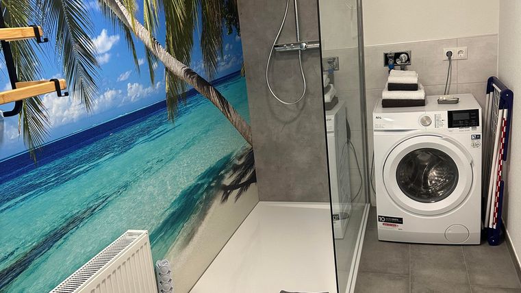 Ein modernes Badezimmer mit einer begehbaren Dusche und einer Waschmaschine. An der Wand ist ein tropisches Strandmotiv mit Palmen zu sehen.