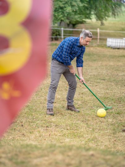 Ein Mann steht auf einer Wiese und spielt mit einem großen gelben Ball. Im Hintergrund ist eine Fahne sichtbar.