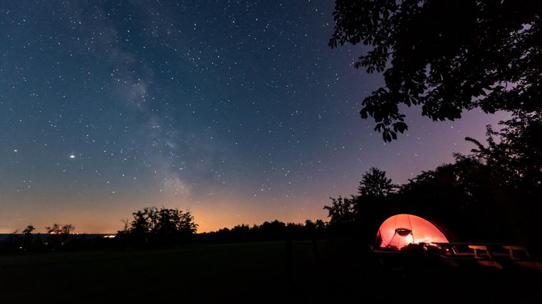 Eine Zeltanlage unter einem sternenklaren Himmel. Die Milchstraße ist sichtbar und das Zelt leuchtet im Dunkeln.