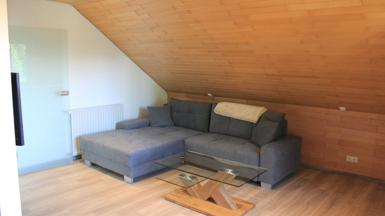 Ein gemütliches Wohnzimmer mit einer grauen Couch und einem Glastisch. Die Wände sind mit Holz verkleidet und der Boden ist aus hellem Holz.
