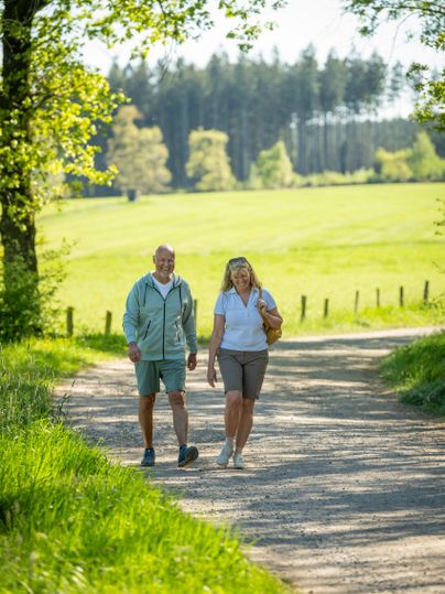 Ein älteres Paar geht auf einem ruhigen Weg durch eine grüne Landschaft. Die Sonne scheint und die Bäume bieten Schatten.