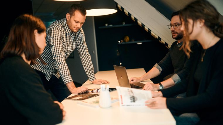 Eine Gruppe von vier Personen sitzt an einem Tisch und diskutiert. Vor ihnen liegen Magazine und ein Laptop.