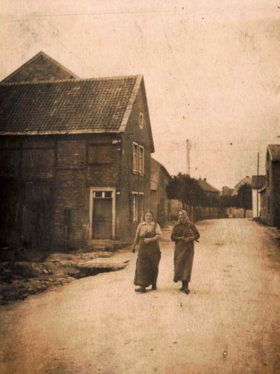 Zwei Frauen gehen eine ruhige Straße entlang, umgeben von alten Gebäuden. Die Szenerie hat einen nostalgischen Charakter und wirkt historisch.