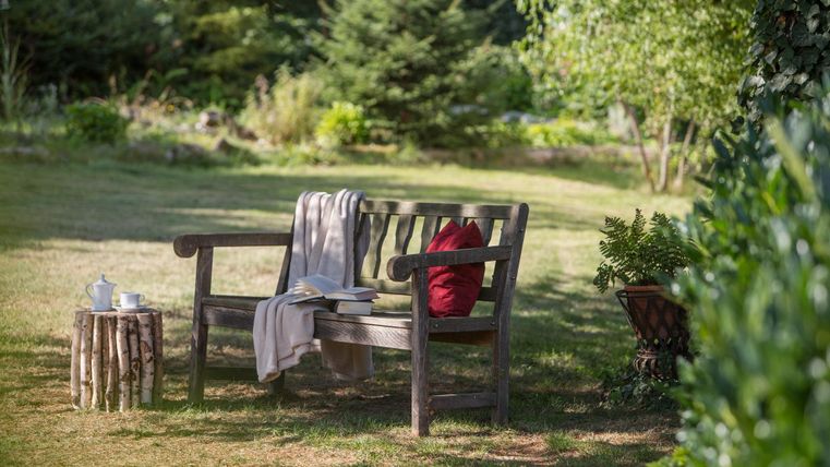 Eine gemütliche Bank im Garten mit einem roten Kissen und einer Decke. Neben der Bank steht ein kleiner Tisch aus Holz.