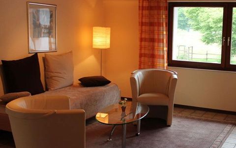 Ein gemütliches Wohnzimmer mit einem Sofa, zwei Sesseln und einem Glastisch. Der Raum hat große Fenster und angenehmes Licht.
