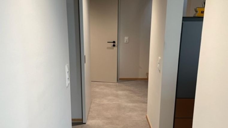 Ein langer Flur mit hellen Wänden und Teppichboden. Am Ende des Flurs befindet sich eine geschlossene Tür.