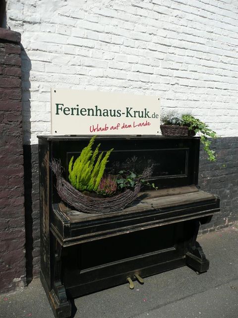 Ein altes Klavier mit Pflanzen dekoriert, steht vor einer weißen Wand. Darüber hängt ein Schild mit der Aufschrift "Ferienhaus-Kruk.de".