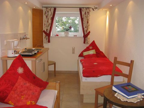 Ein gemütliches Zimmer mit zwei Einzelbetten und roten Kissen. Der Raum hat eine kleine Küchenzeile und ein Fenster mit Blick ins Freie.