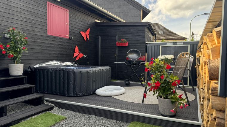 Ein gemütlicher Außenbereich mit einem aufblasbaren Whirlpool, Pflanzen und roten Schmetterlingen. Eine einladende Terrasse mit einem Grill und Sitzgelegenheiten.