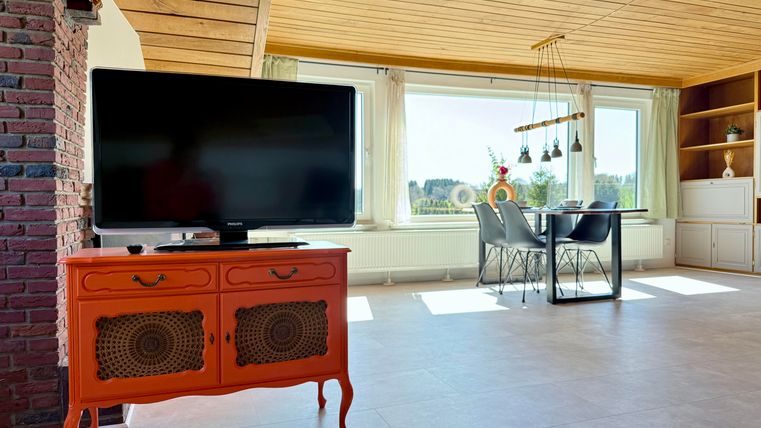 Ein modernes Wohnzimmer mit einem orangefarbigen Sideboard und einem großen Fernseher. Der Raum hat große Fenster mit Blick ins Grüne und einen Esstisch mit Stühlen.