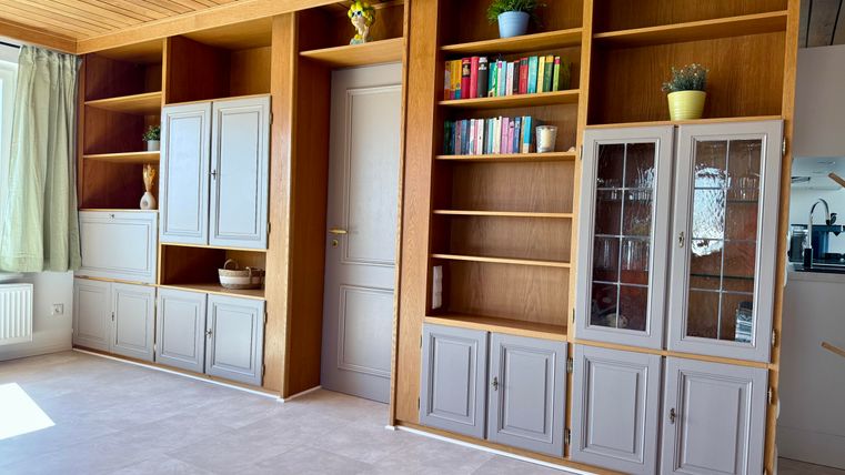 Ein heller Raum mit modernen, integrierten Regalen und einem Holzdecke. Die Regale sind teils geschlossen und teils offen und beherbergen Bücher und Dekorationselemente.