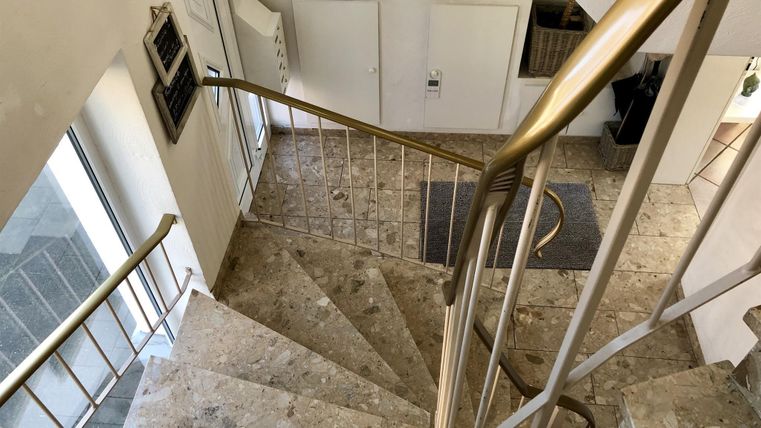 Eine elegante Treppe aus Stein mit einem gelbgoldenen Geländer. Der Raum ist hell und modern gestaltet mit großen Fenstern.