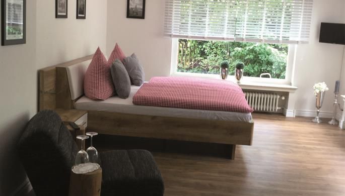 Ein modernes Schlafzimmer mit einem gemütlichen Bett und karierten Kissen. Große Fenster sorgen für viel Tageslicht und einen Blick ins Grüne.