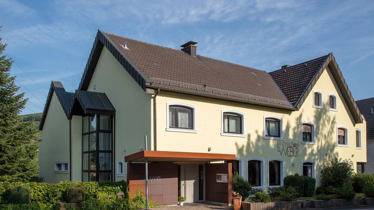 Ein modernes Gebäude mit gelber Fassade und einem Schattendach. Umgeben von Bäumen und einem gepflegten Garten.